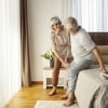 Helpful old woman is helping her ill husband to stand up from a bed in the morning.