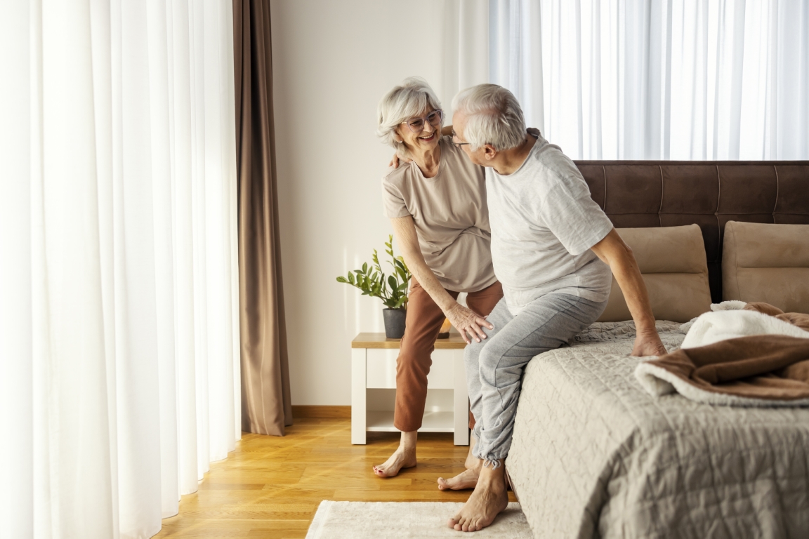 Helpful old woman is helping her ill husband to stand up from a bed in the morning.
