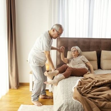 A caring old man is helping his ill wife to stand up from a bed in the morning.