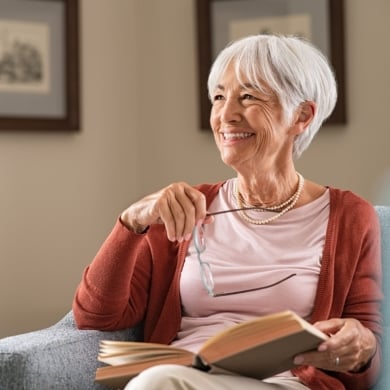 Cheerful senior woman holding book and eyeglasses thinking while relaxing at home. Happy elderly woman reading book at home sitting on couch. Beautiful old teacher takes a break from reading while looking through the window with a big grin.