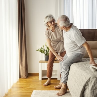 Helpful old woman is helping her ill husband to stand up from a bed in the morning.